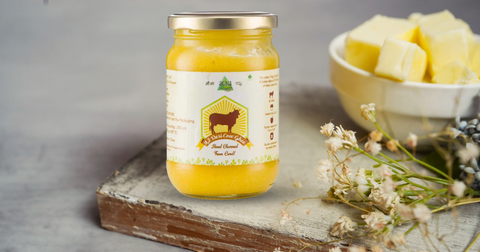 A glass jar of golden ghee placed on a rustic wooden surface, with fresh butter in the background.